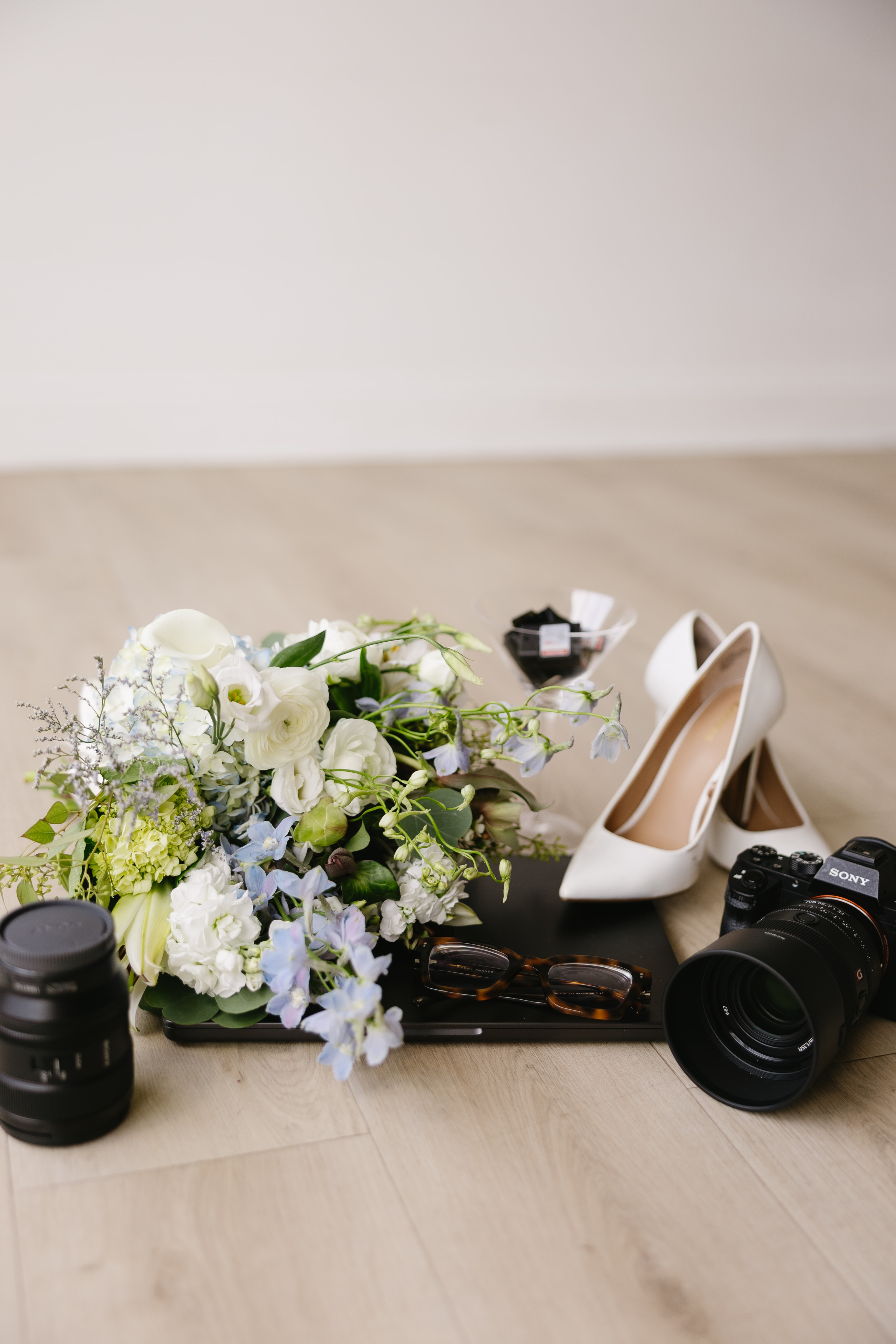 Styled flat lay with a pastel bouquet, white heels, glasses, and a camera on wood flooring.