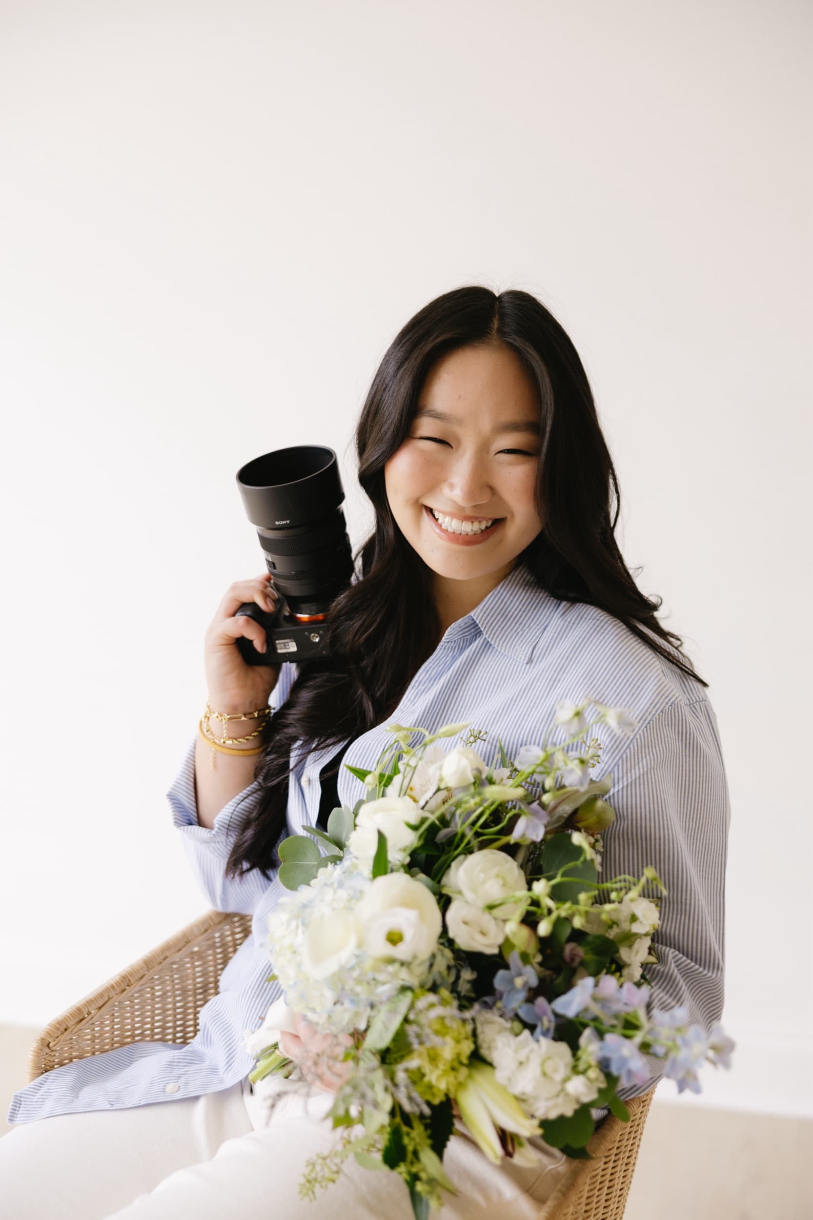 Photographer smiling while holding a camera and a bouquet of white and blue flowers.