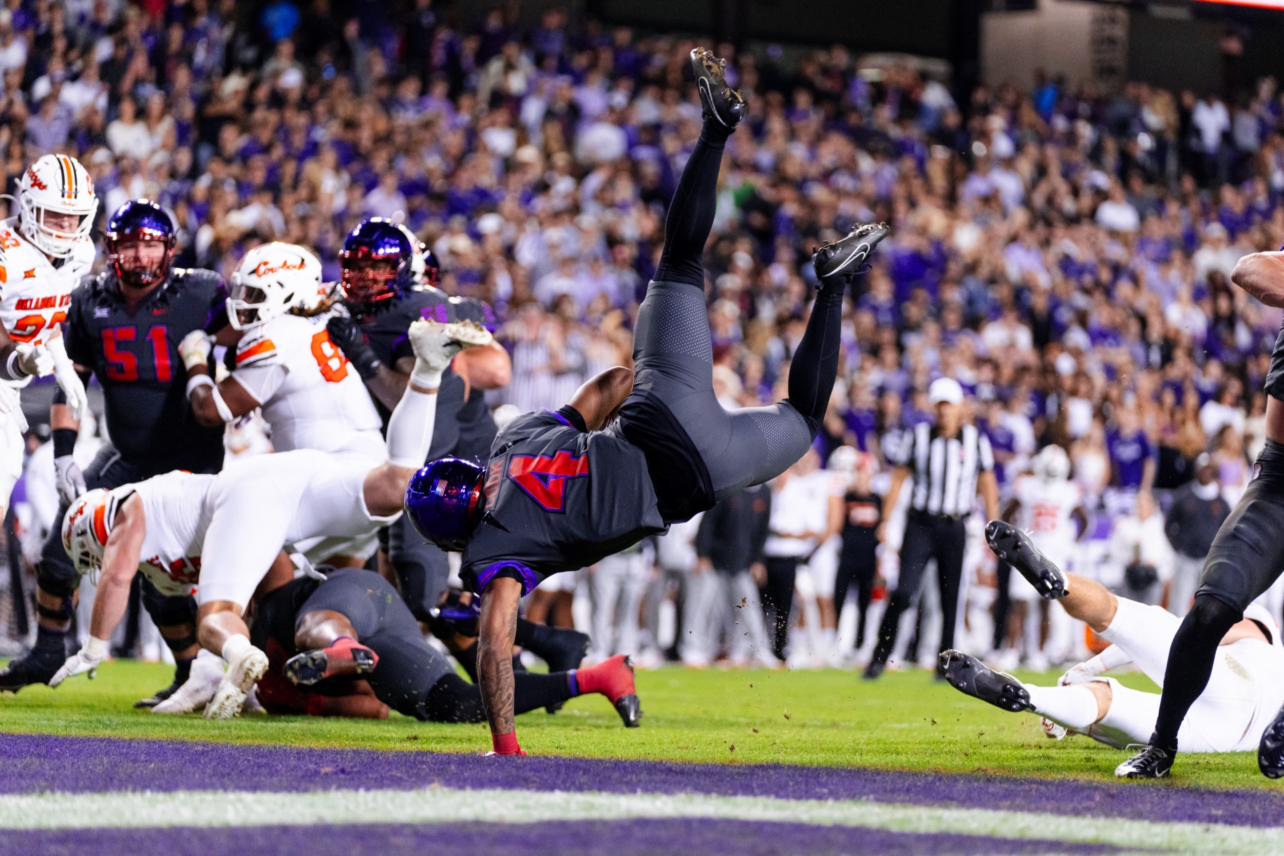 Football player flipping upside down mid-air, diving toward the end zone under stadium lights.