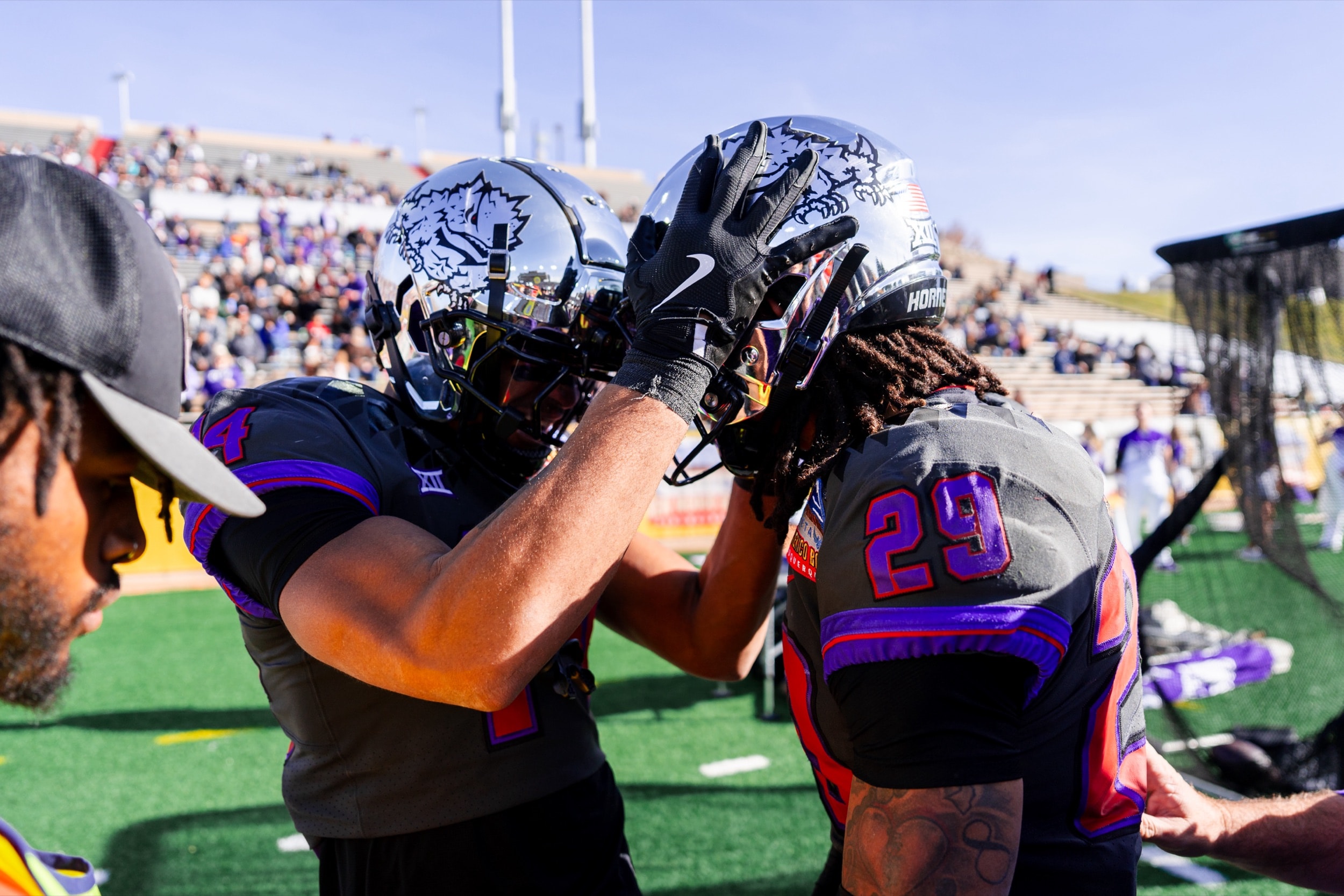 Football teammates celebrating on the sideline, patting each other's helmets under a clear sky.
