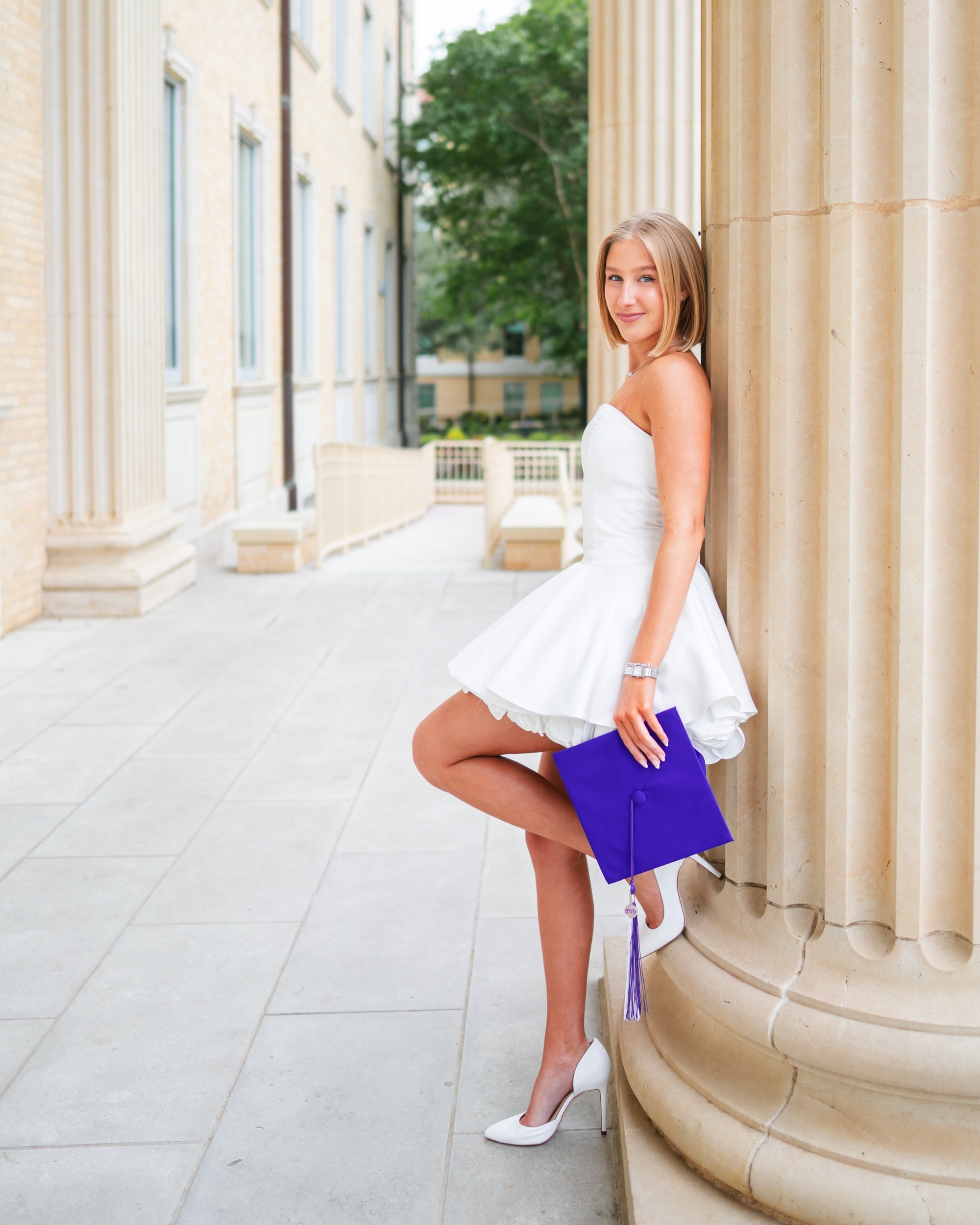Graduate in a white dress leaning against a stone column, holding her graduation cap.
