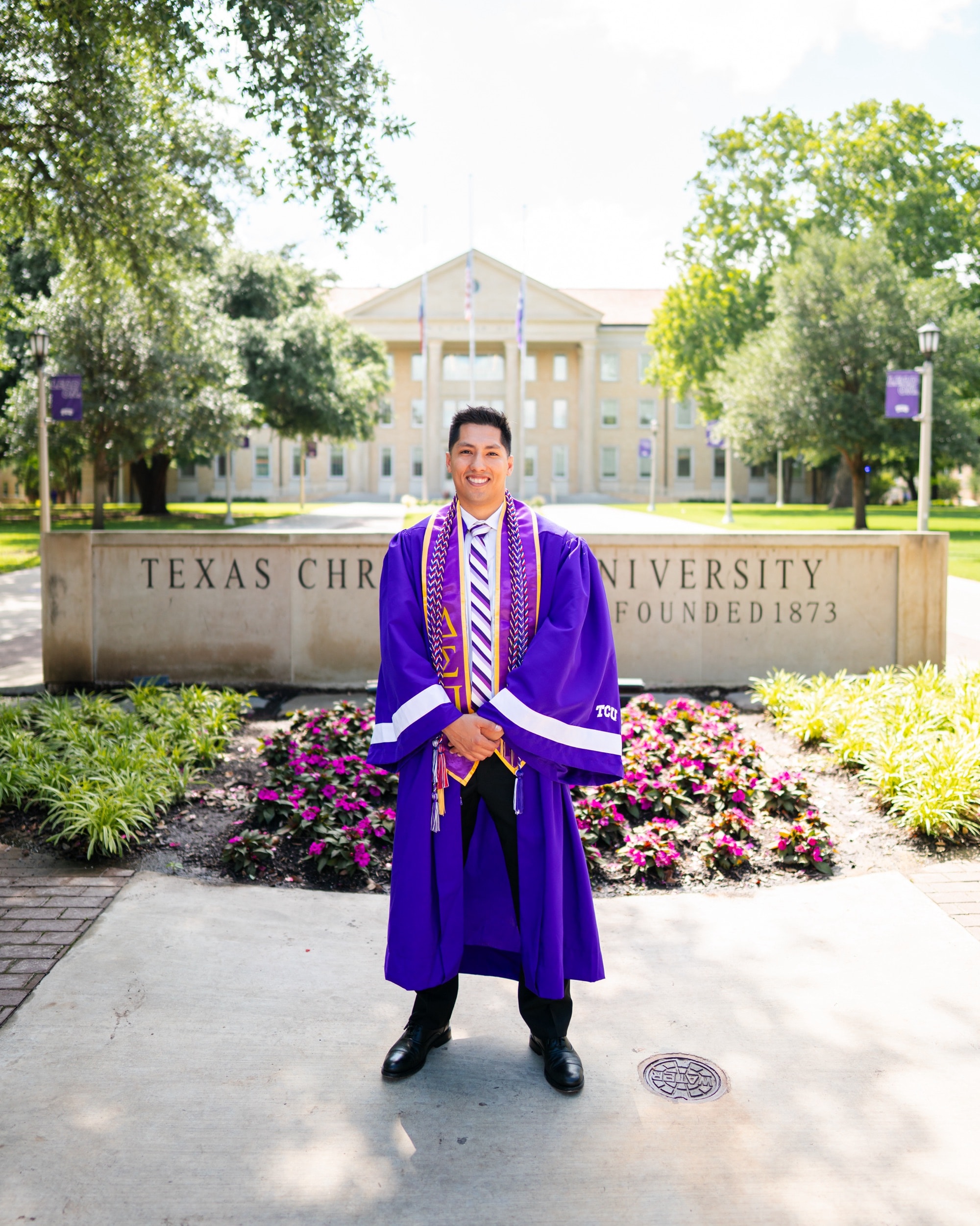 Graduate in a purple gown and honor cords standing proudly before a university sign.