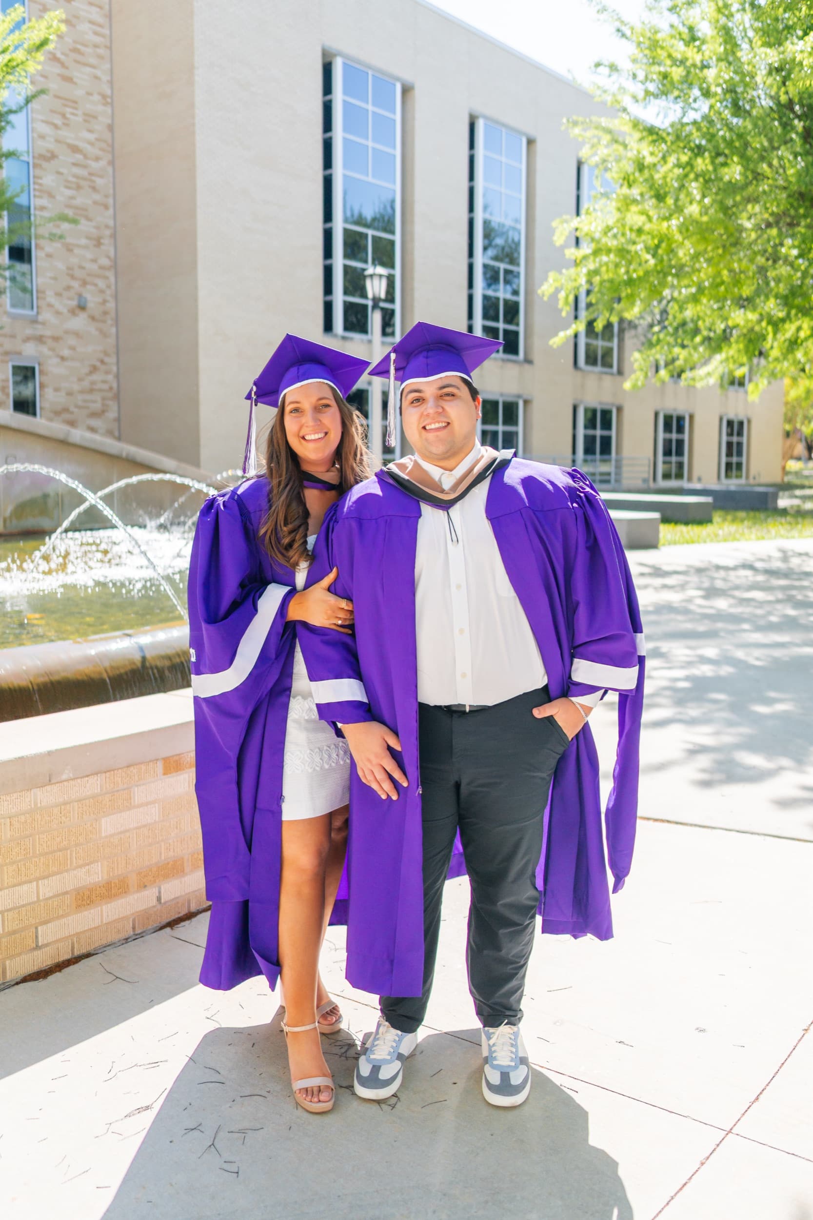 Graduating couple in purple caps and gowns embracing warmly beside a sunlit campus fountain.