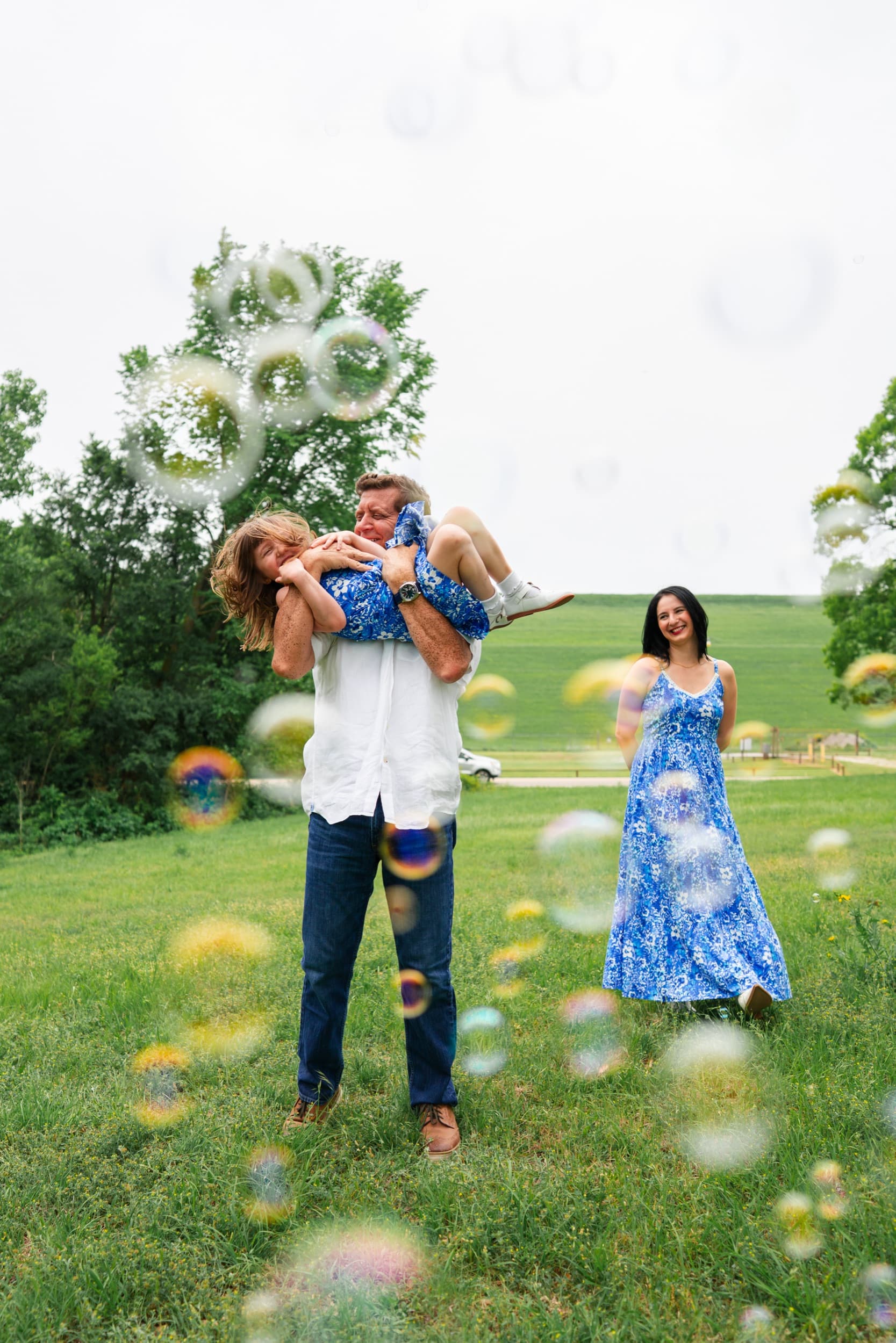 Father joyfully lifting a laughing child as mom watches, bubbles drifting through a grassy field.