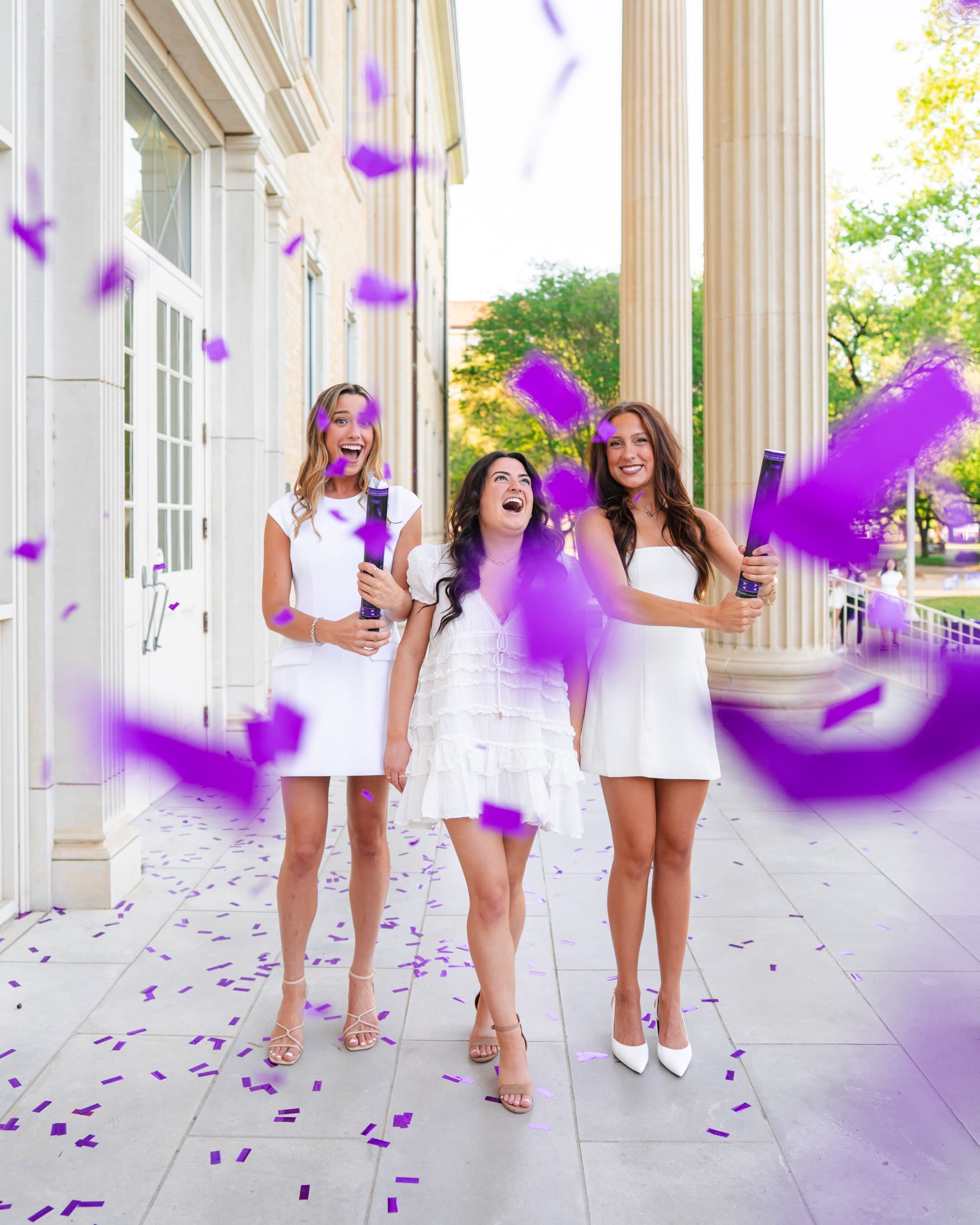 Three friends in white dresses laughing as purple confetti bursts around them beside a campus building.