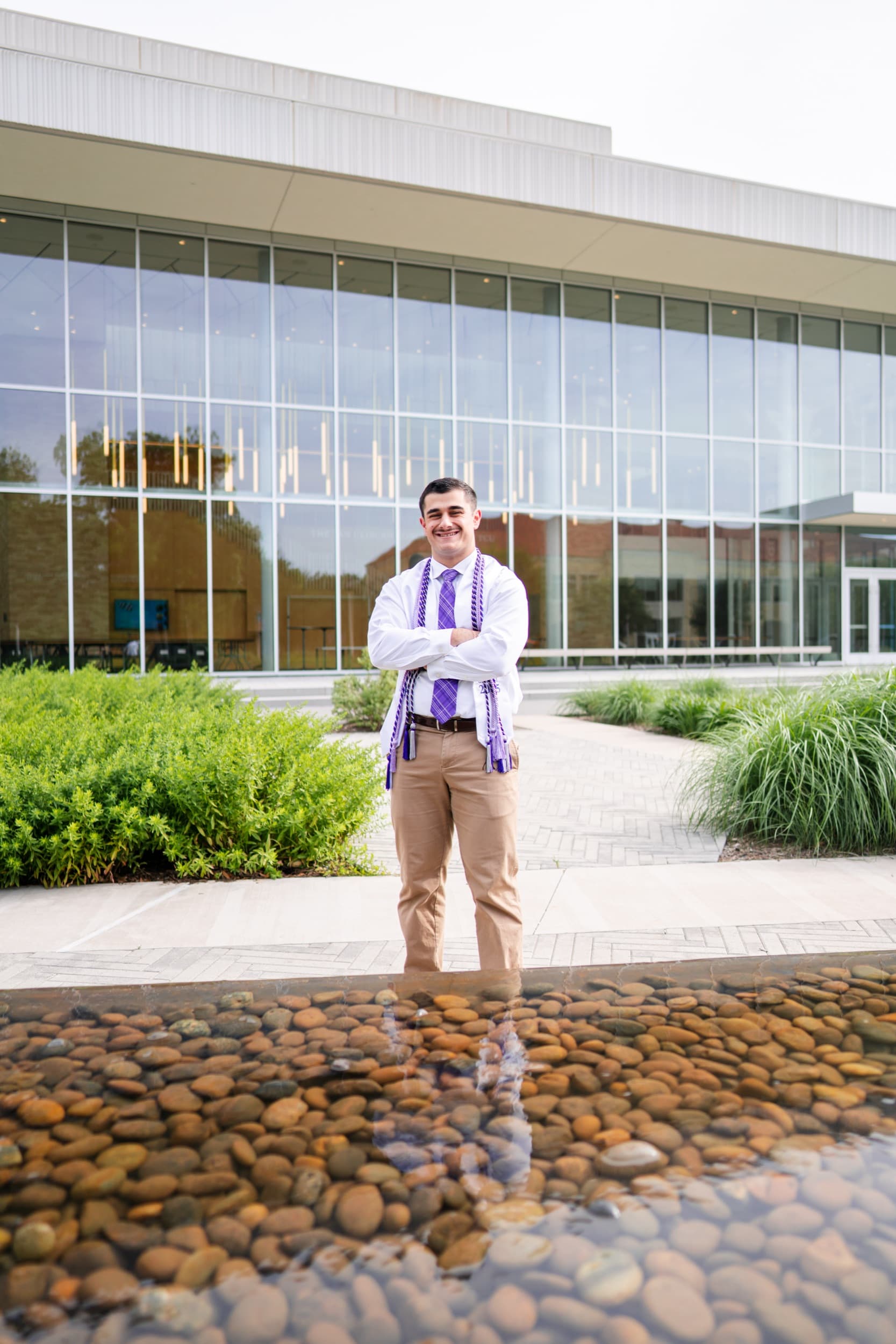 Smiling graduate wearing purple honor cords stands by a reflecting pool outside a glass building.