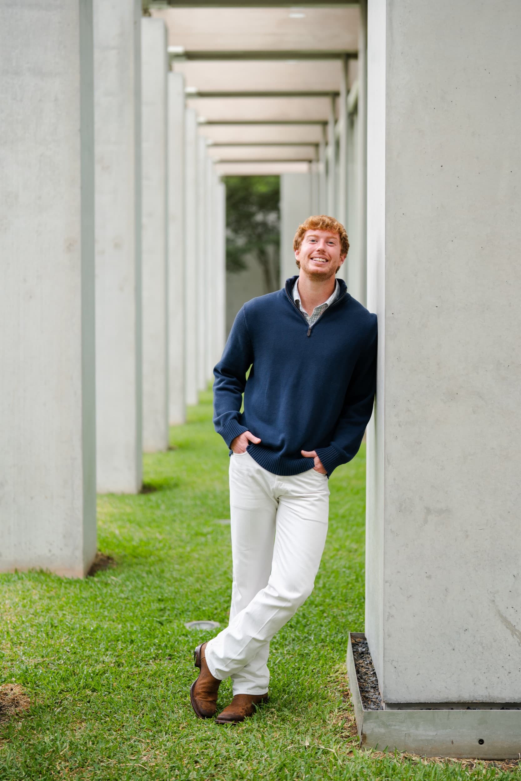 Senior in a navy quarter-zip leaning against a concrete column in a shaded outdoor colonnade.