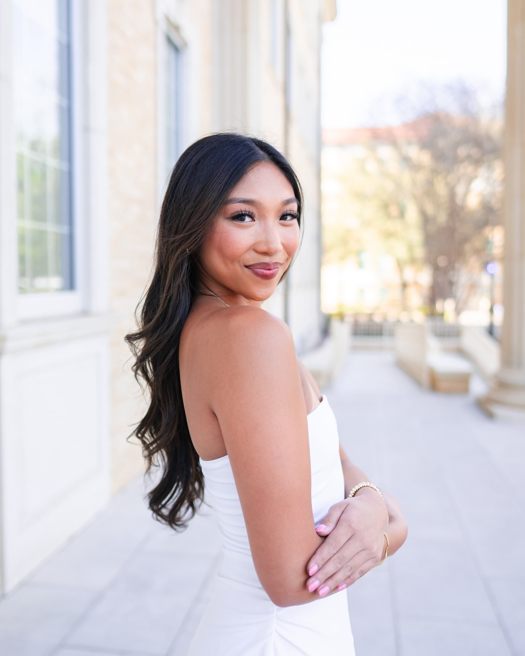 Senior in a white strapless dress glancing back over her shoulder along a sunlit colonnade.